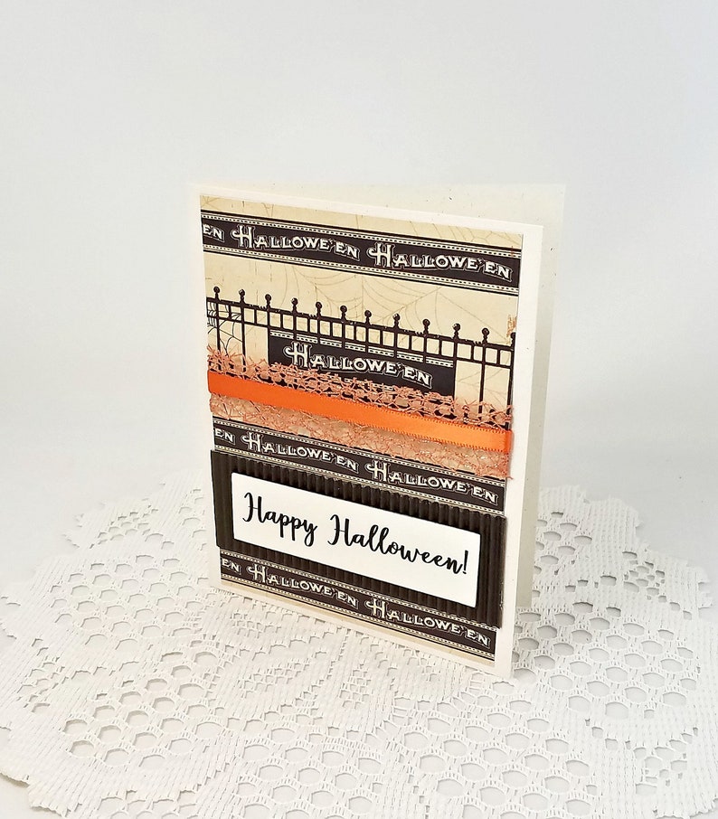 May include: A handmade Halloween greeting card with a cream-colored background. The card features decorative black and orange ribbons, a black fence design, and the words "Happy Halloween!" in white text.