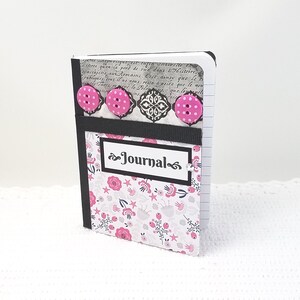 May include: A small, black and white journal with a floral pattern and pink buttons. The journal has a black ribbon and the word "Journal" written on the front.