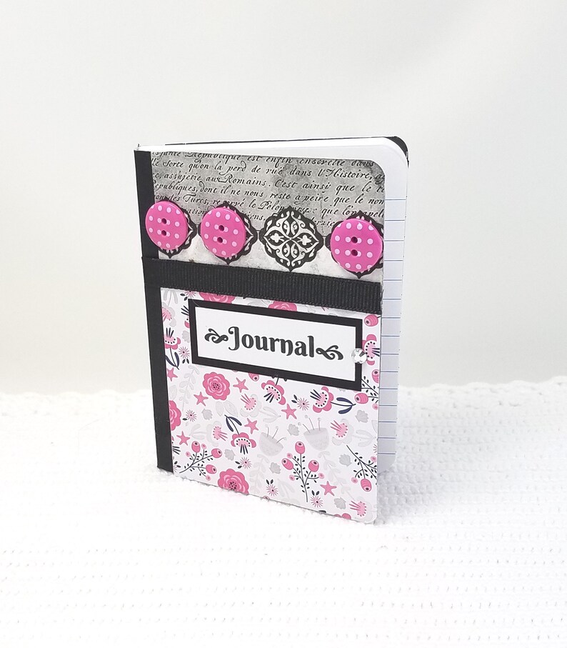 May include: A small, black and white journal with a floral pattern and pink buttons. The journal has a black ribbon and the word "Journal" written on the front.