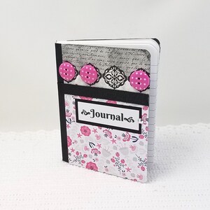 May include: A small black and white journal with a floral design and pink buttons. The journal has a black ribbon and a white label that says "Journal".