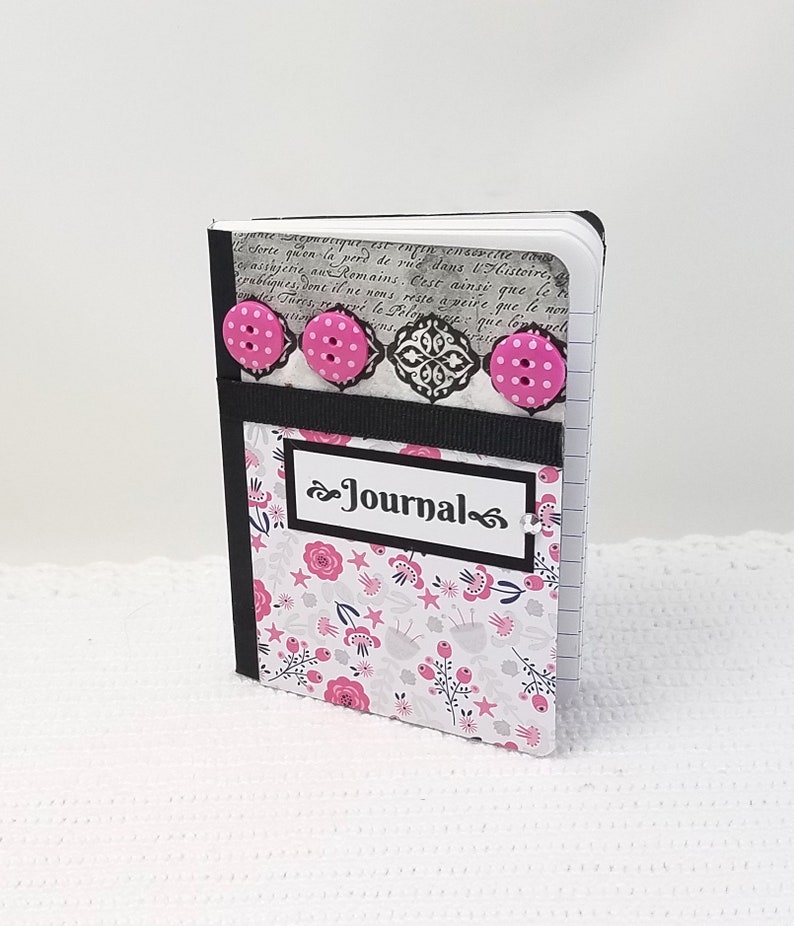 May include: A small black and white journal with a floral design and pink buttons. The journal has a black ribbon and a white label that says "Journal".