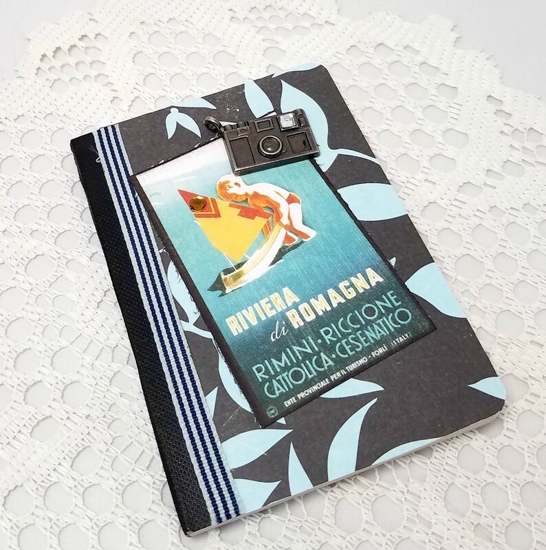 Puede incluir: Una libreta negra y azul con un p&oacute;ster de viaje vintage en la portada. El p&oacute;ster presenta a un ni&ntilde;o en un velero con el texto "Riviera di Romagna Rimini-Riccione Cattolica-Cesenatico" y un peque&ntilde;o amuleto de c&aacute;mara plateado.