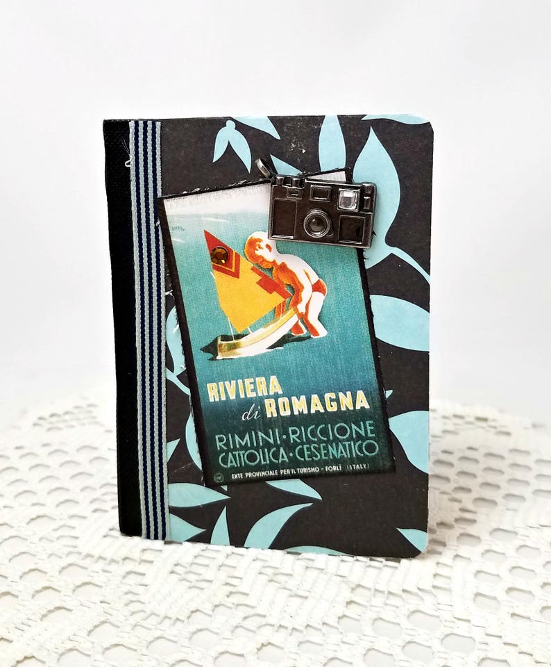 Puede incluir: Un diario de viaje negro y azul con un amuleto de c&aacute;mara vintage y un p&oacute;ster de viaje vintage en la parte delantera. El p&oacute;ster presenta a un ni&ntilde;o navegando en un velero frente a un oc&eacute;ano azul y turquesa. El texto en el p&oacute;ster dice "Riviera di Romagna Rimini-Riccione Cattolica-Cesenatico Ente Provinciale per il Turismo - Forli (Italy)"
