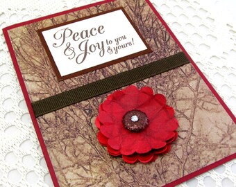 Puede incluir: Una tarjeta roja y marrón con una flor roja y el texto "Peace & Joy to you & yours!" sobre un fondo blanco.
