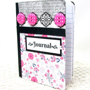 May include: A small, black and white journal with a floral pattern and pink buttons. The front cover has a black label with the word "Journal" written in a decorative font.