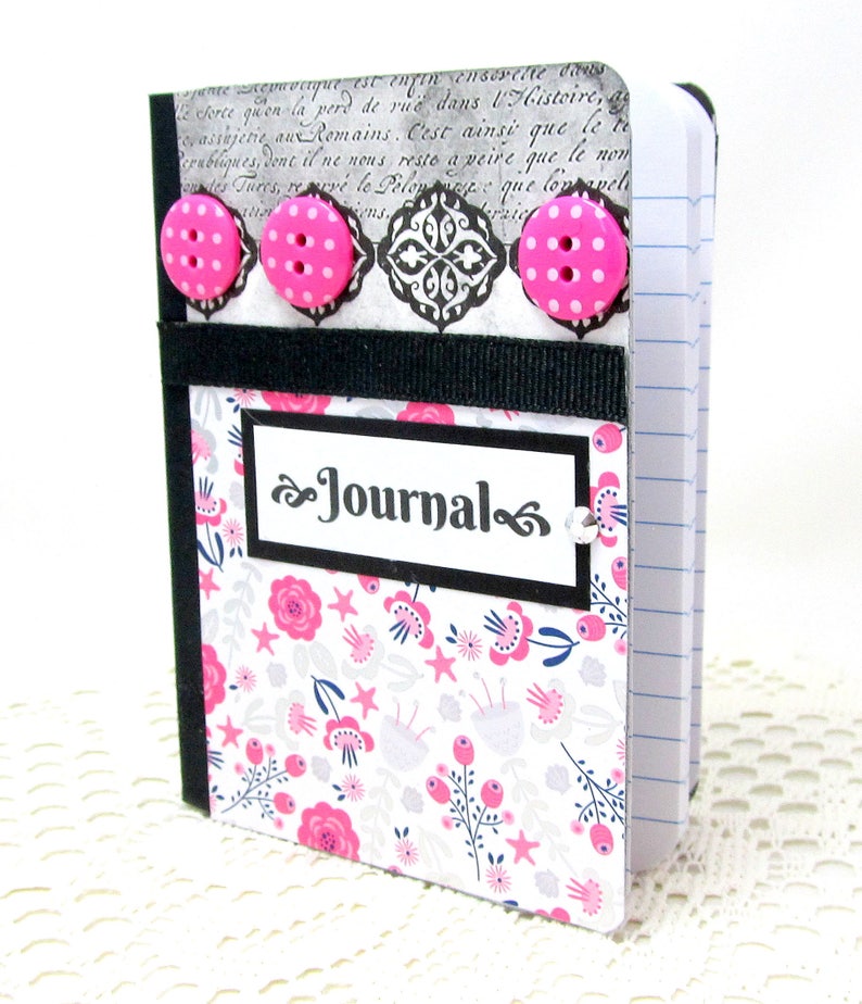 May include: A small, black and white journal with a floral pattern and pink buttons. The front cover has a black label with the word "Journal" written in a decorative font.