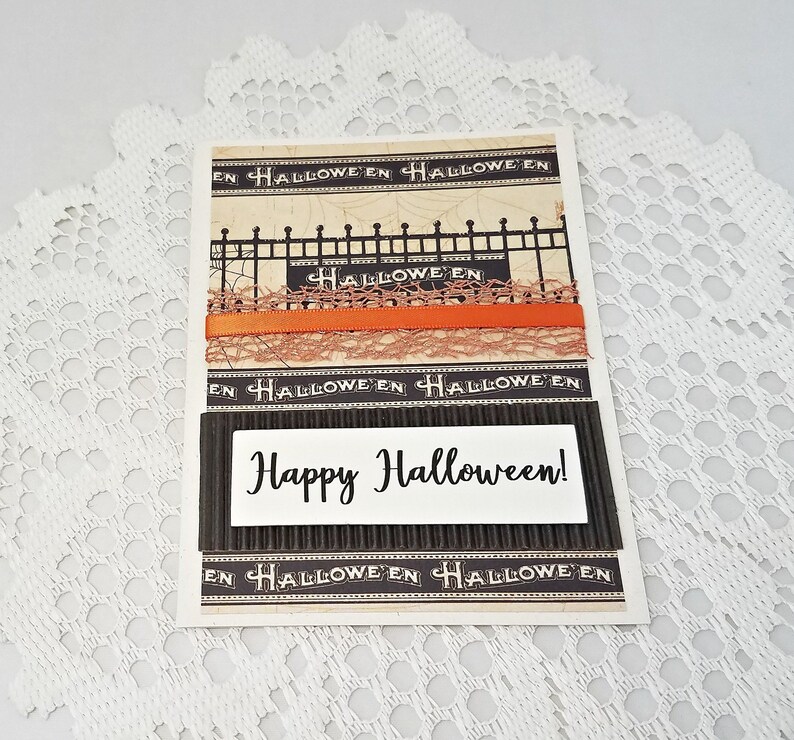 May include: A Halloween greeting card with a decorative design. The card features the text "Happy Halloween!" on a white rectangle, with orange ribbon and lace accents. The background has a black and beige color scheme.