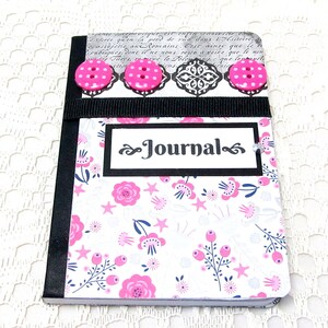 May include: A small, white journal with a black border and a floral pattern. The cover has a black ribbon and three pink buttons. The word "Journal" is written in black on a white background.
