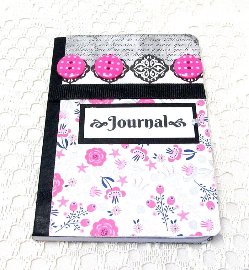 May include: A small, white journal with a black border and a floral pattern. The cover has a black ribbon and three pink buttons. The word "Journal" is written in black on a white background.
