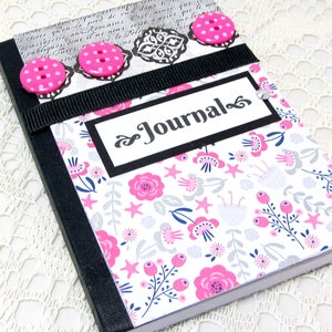 May include: A black journal with a floral design cover. The cover has a black ribbon and pink buttons. The title "Journal" is written in black cursive on a white background.