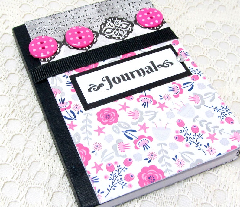 May include: A black journal with a floral design cover. The cover has a black ribbon and pink buttons. The title "Journal" is written in black cursive on a white background.