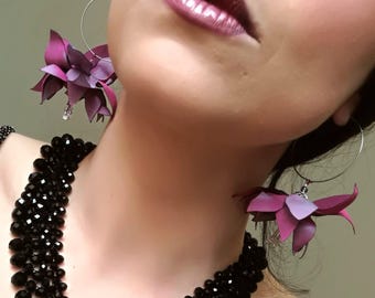 Puede incluir: Un par de pendientes de aro de plata con detalles de flores moradas. Los pendientes los lleva una persona con el pelo oscuro y un collar de cuentas negras.