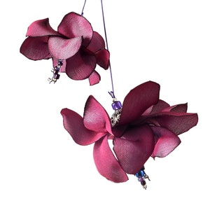 Könnte beinhalten: Zwei burgunderfarbene Stoffblumen mit violetten und silbernen Perlen, die an Fäden hängen. Die Blumen sind aus Stoff gefertigt und haben einen zarten, femininen Look.