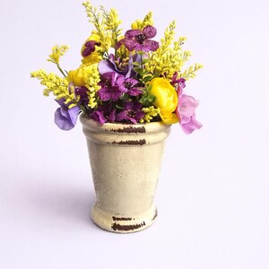 May include: A floral arrangement in a cream-colored, distressed ceramic vase. The bouquet features yellow, purple, and pink flowers, including tulips and other blooms. The vase has a weathered, aged appearance, adding a rustic touch to the display.