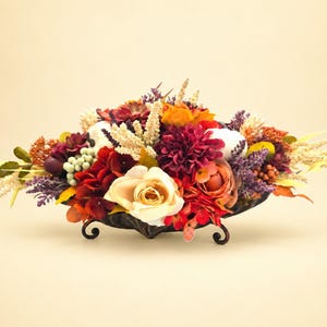 May include: A floral arrangement in a black metal bowl featuring a mix of cream, deep red, and orange flowers, including roses and dahlias, accented with white sprigs, green berries, and purple filler flowers, creating a dense, colorful display.