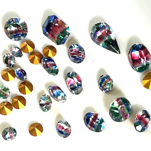 Peut inclure: Un assortiment de pierres précieuses irisées et de strass dorés à dos plat est dispersé sur une surface blanche. Les pierres précieuses présentent des teintes roses, bleues et vertes. Les strass sont ronds et ont une finition métallique.