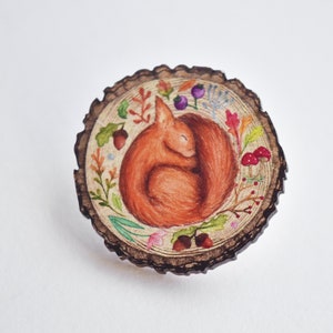 May include: A wooden pin with a painted design of a curled up squirrel sleeping in a forest scene. The squirrel is brown and the scene is surrounded by green leaves, red berries, and brown acorns.