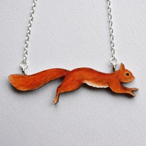 Könnte beinhalten: Halskette mit silberner Kette und Anhänger in Form eines rennenden Eichhörnchens. Das Eichhörnchen ist in Orange- und Brauntönen bemalt, mit detailliertem Fell. Ein naturbezogenes Accessoire.