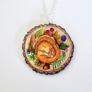 May include: A wooden pendant necklace with a painted image of a sleeping dormouse surrounded by flowers, leaves, and berries. The pendant is shaped like a tree trunk slice.