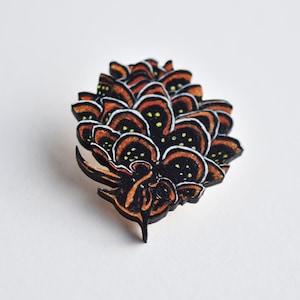 Puede incluir: Un pin de madera con forma de piña. El cono está pintado con detalles negros, naranjas y blancos. El pin es una excelente manera de agregar un toque de naturaleza a tu estilo.