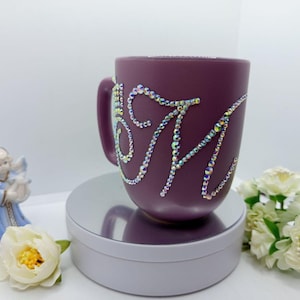 Pode incluir: Uma caneca de cerâmica cor de ameixa adornada com strass brilhantes formando as letras "S M". A caneca está sobre uma plataforma giratória branca, com uma pequena figura de anjo e flores brancas no fundo. Um item decorativo.