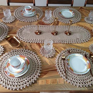 May include: A beige crochet table setting featuring round placemats, matching coasters, and an oval table runner. The set includes white ceramic plates and cups with orange and brown rims, clear textured glass tumblers, and gold-toned metal candlesticks.