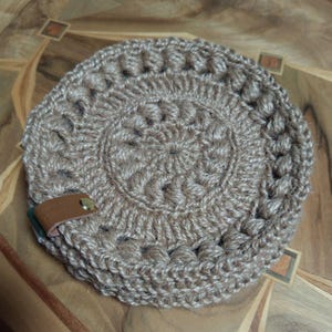 May include: A round, tan, crocheted coaster made from textured jute yarn. The design features a central spiral pattern surrounded by a ring of raised puff stitches. A small, folded brown leather tab is attached to the edge with a gold metal rivet.