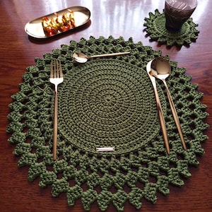 Puede incluir: Un mantel individual de ganchillo verde oliva hecho a mano con un posavasos a juego. Tenedor, cuchara y cuchillo dorados están colocados sobre el mantel individual. Una pequeña bandeja dorada con caramelos y un vaso de agua completan la mesa.