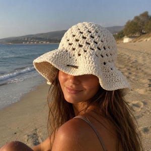 Pode incluir: Um chapéu de crochê estilo bucket hat na cor creme, com um padrão de trama aberta e uma aba larga e flexível. Uma pequena etiqueta retangular de metal dourado está fixada na parte central frontal do chapéu, logo acima da aba, como detalhe.