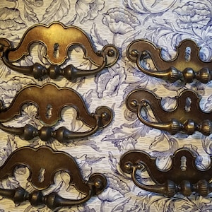 May include: Six antique bronze-toned drawer pulls with keyhole details and decorative handles. The pulls are arranged on a floral patterned background, showcasing their ornate design and vintage aesthetic. Ideal for furniture restoration projects.