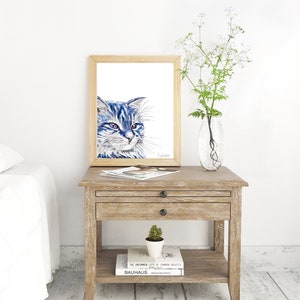 Puede incluir: Una pintura de acuarela enmarcada de un gato azul con ojos verdes. El gato est&aacute; mirando hacia la derecha del marco. La pintura est&aacute; en una mesita de noche de madera con un libro y una peque&ntilde;a planta en maceta.