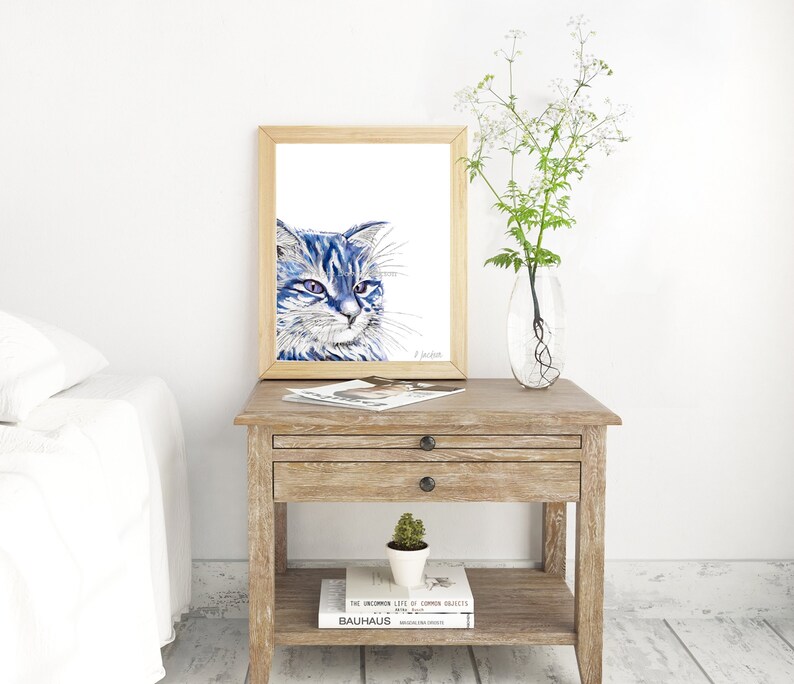 Puede incluir: Una pintura de acuarela enmarcada de un gato azul con ojos verdes. El gato est&aacute; mirando hacia la derecha del marco. La pintura est&aacute; en una mesita de noche de madera con un libro y una peque&ntilde;a planta en maceta.