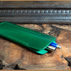 May include: A green leather pen case with two silver pens inside. The case is stitched with white thread and has a smooth, glossy finish.