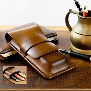 May include: A brown leather pen case with a flap closure. The case is sitting on a wooden table next to a brass pitcher filled with pens.