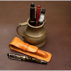 May include: A brown leather pen case with a single pen inside. The pen has a black and brown marbled design. The case is sitting on a brown surface next to a brass pitcher with three pens inside. The pens are black, red, and white.