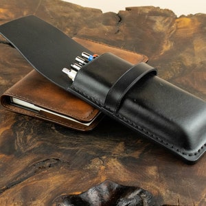 May include: A black leather pen case with a flap closure and a brown leather notebook underneath. The pen case has a stitched design and is closed with a strap.