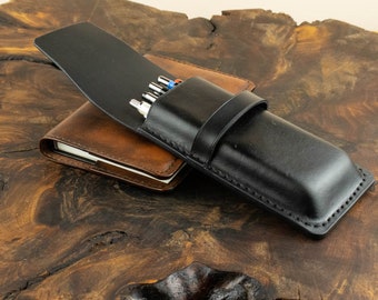 May include: A black leather pen case with a flap closure and a brown leather notebook underneath. The pen case has a stitched design and is closed with a strap.