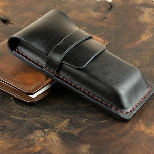 Op de afbeelding: Een zwart leren pennenetui met rode stiksels en een klep sluiting. De etui ligt op een houten oppervlak.