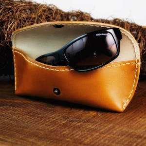 May include: A brown leather sunglasses case with a white interior and a pair of black sunglasses with the word "POLARIZED" printed on the lens.