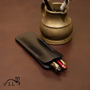 Puede incluir: Un estuche de cuero negro para bolígrafos con costuras moradas, que contiene dos bolígrafos con detalles dorados. Un bolígrafo es rojo y el otro es dorado.