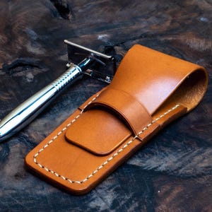 May include: A brown leather case with white stitching for a safety razor. The case has a flap that folds over to protect the razor.