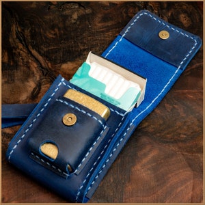 Puede incluir: Un estuche de cigarrillos de cuero azul con costuras blancas y un cierre de botón a presión de latón. El estuche contiene un encendedor dorado y un paquete de cigarrillos. El estuche está abierto, revelando el contenido. El fondo es una superficie de madera oscura.