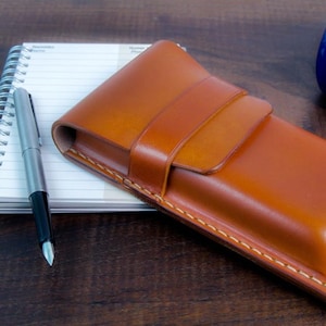 Puede incluir: Un estuche para bolígrafos de cuero marrón con cierre de solapa y detalles de costura. El estuche está sobre un cuaderno con un bolígrafo plateado.