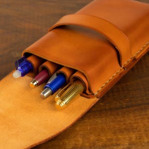 Leather pencil case in sadle tan , with compartments for 4 pens lying on a wooden board