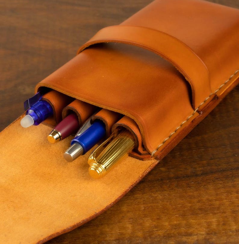 Leather pencil case in sadle tan , with compartments for 4 pens lying on a wooden board