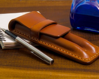 May include: A brown leather pen case with two slots for pens. The case is closed and a silver pen is lying next to it on a wooden surface.