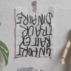 Könnte beinhalten: Ein Blatt Papier mit schwarzem Text ist mit gestreiftem Klebeband an einer weißen Wand befestigt. Der Text lautet "UNPORTRAIT EX TRAOR DINAIRE". Eine Vintage-Kamera und eine Holzpuppe befinden sich im Vordergrund.