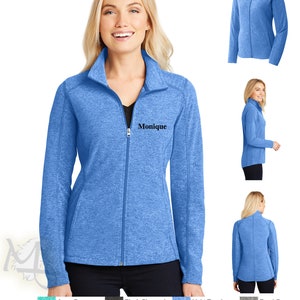 Puede incluir: Una chaqueta azul claro con cremallera y cuello alto. La chaqueta tiene un pequeño nombre bordado "Monique" en el pecho izquierdo. La chaqueta se muestra en una modelo que lleva pantalones negros.