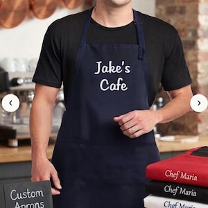 May include: A navy blue apron featuring the white embroidered text "Jake's Cafe" on the chest. A stack of folded aprons in red, black, white, and blue sits nearby, each personalized with white text reading "Chef Maria" or "Baker's Dozen".