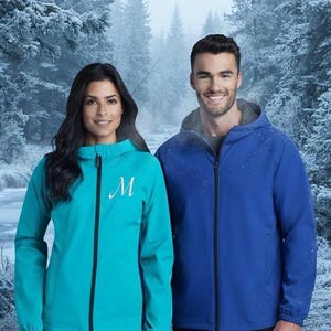 Op de afbeelding: Een turquoise jack met capuchon, zwarte ritssluiting en een witte letter M op de borst, naast een blauw jack met capuchon en zwarte ritssluiting. Beide jacks hebben lange mouwen, elastische boorden en een gladde, waterafstotende textuur.